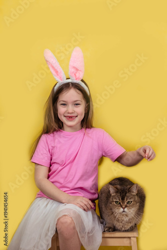 Young girl wearing pink bunny ears and a pink shirt sits on a stool beside a fluffy cat against a bright yellow background