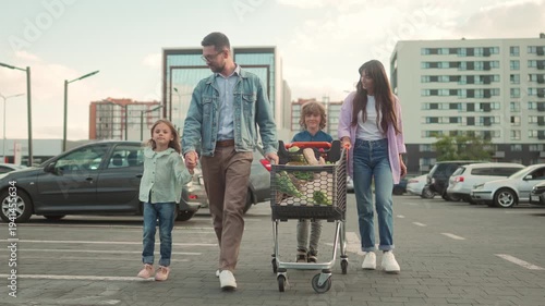 Family walking across parking lot after shopping. Father holding daughter’s hand, son pushing cart full of groceries, mother smiling beside. Parents and children spending time together outdoors.