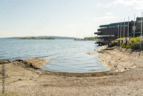 View of the serene, rocky shoreline meets the tranquil waters of the fjord, contrasted by a modern building under a clear sky, Oslo, Oslo, Norway.