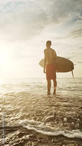 Young surfer stands on the beach at sunset, holding his surfboard near a majestic cliff. Warm backlight from the sun illuminates the scene, with sparkling ocean waves in the background, creating a