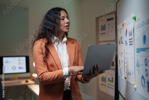 Businesswoman presenting financial charts and graphs on a board while holding a laptop in an office, focused on strategy, analytics and growth planning for corporate success