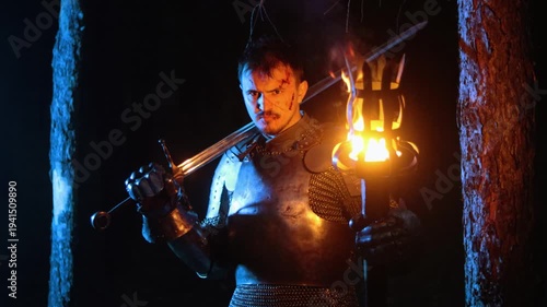 A knight in full armor, holding a sword and a torch, stands in a dark night forest. A medieval warrior with a bloodied face searches in the darkness.
