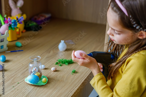 Little girl using a hot glue gun to decorate colorful easter eggs for a creative DIY holiday craft project at home