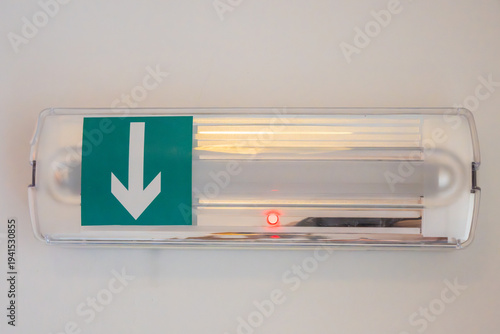 Принти на полотні an illuminated emergency exit light fixture mounted on a plain white wall, featuring a green directional sign with a white downward arrow and a small red LED indicator