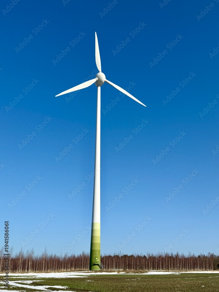 Fototapeta premium Single wind turbine standing in an open field with forest line in the background under clear blue sky and bright daylight.