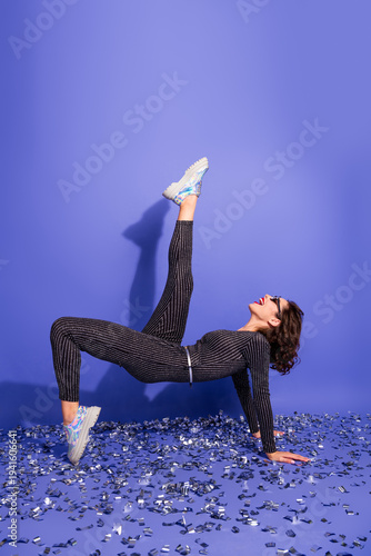Foto Young chic woman in shiny striped jumpsuit performs fitness pose on blue studio