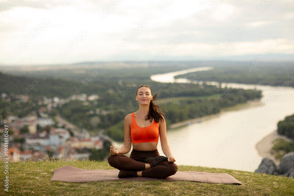 Fototapeta premium Lady sitting on mat, meditating and practicing yoga beside river surrounded by hills and city in the background. Mindful mountain escape