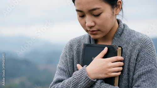 Contemplative East Asian woman in grey cardigan holds a black Bible to her chest against a blurred outdoor background, to enhance spiritual content.
