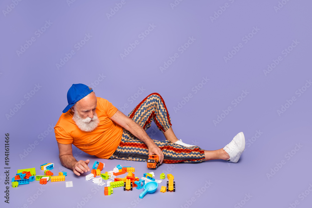 Fototapeta premium Older man in orange tshirt and striped pants plays with colorful blocks on a purple violet background