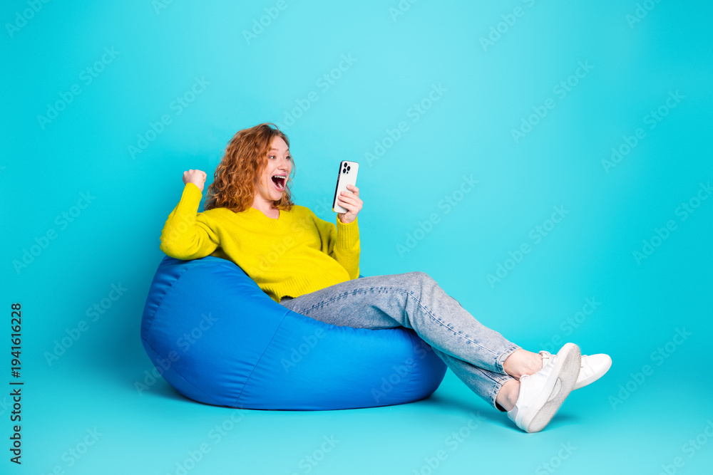 Obraz premium Young woman in a bright yellow sweater relaxing on a blue beanbag smiles at her phone in a vibrant teal studio background