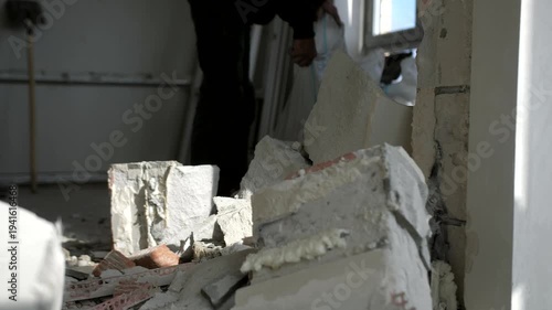 Man wearing gloves gathers broken concrete pieces into trash bag after wall demolition inside apartment renovation site.