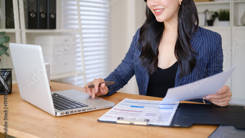 Close up of businesswoman reviewing financial documents while using a laptop at a modern office desk.