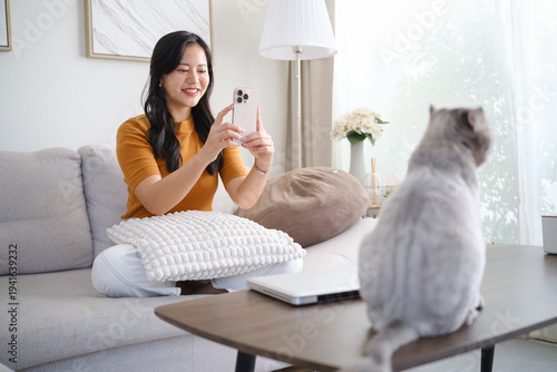 Young Asian woman taking a photo of her cat with a smartphone while relaxing on a sofa at home.
