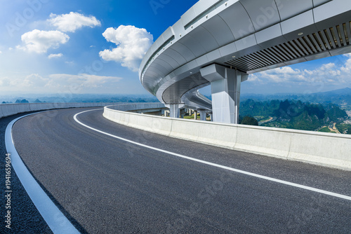 Curving asphalt highway and modern overpass bridge spanning a green mountain valley under a clear blue sky; transportation infrastructure concept.