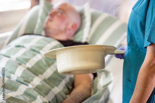 Healthcare worker holding a bedpan for patient hygiene while a male patient lies in hospital bed during nursing care