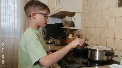 Konstfotografi Young boy wearing eyeglasses standing by an induction stove, carefully preparing
