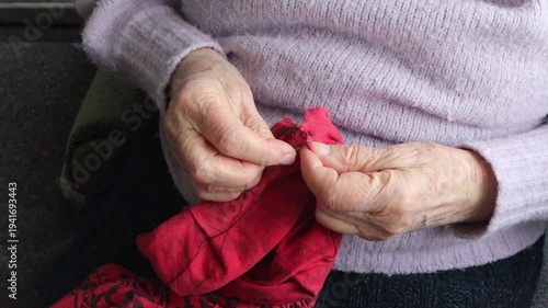 The wrinkled hands of elderly people carefully stitch red fabric with a needle and thread.