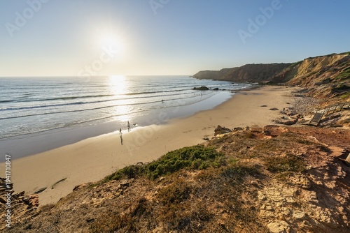 Sunset at  Praia do Amado Costa Vicentina beach, Algarve, Portugal. Golden hour view of a serene beach with gentle waves, rocky cliffs, and people walking along the shore in southern Portugal.