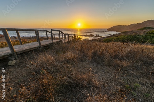 Sunset over Costa Vicentina, Algarve, Portugal with wooden boardwalk leading to the sea. Golden sunset over the ocean with wooden walkway leading to beach, dry grass foreground, rocky coastline