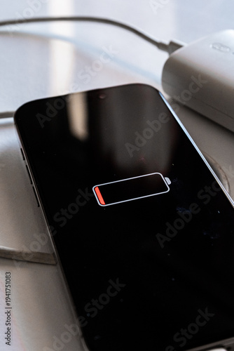 A smartphone with a low battery icon is connected to a white power bank via a charging cable. The scene is set on a reflective surface.