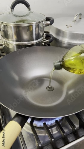 Vertical video social media - Closeup of cooking oil being poured from a plastic bottle into a new wok which is heating up over a kitchen cooker hob flame, with other pots and pans behind.