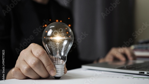 Businessman holding light bulb with glowing gear mechanism over office desk and laptop. Concept of innovation, creative idea, problem solving, technology strategy, business solution 