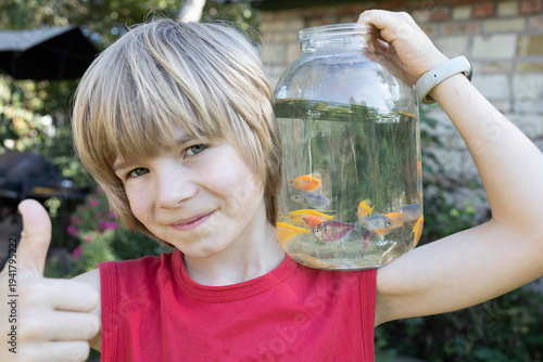 boy holds a transparent jar with small aquarium fish floating in it on his shoulder. Buying colorful fish for an aquarium, a child getting interested in the world of fish, a hobby of aquarium keeping