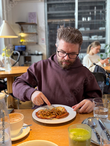 Man enjoying potato pancakes and mushroom sauce at wooden cafe table. Everyday restaurant dining, urban lifestyle routine, comfort food culture and relaxed social atmosphere.