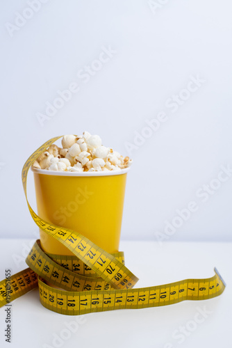 Yellow cup of popcorn wrapped with measuring tape. Dietary сontrol and healthy eating concept.