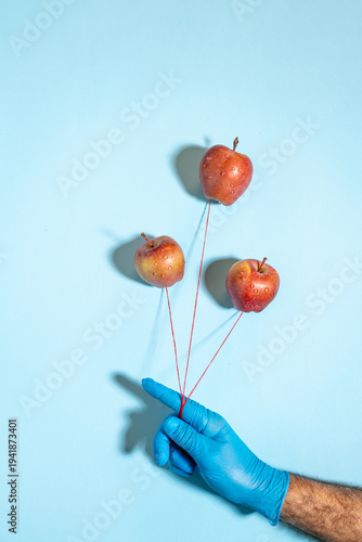Creative concept of hand in blue glove holding red apples as floating balloons on strings against blue background.