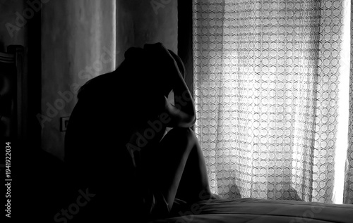 Silhouette of depressed young woman sitting in dark bedroom.  Concept of disappointment, thinking, loneliness, depression.