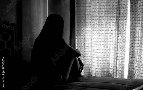 Silhouette of depressed young woman sitting in dark bedroom.  Concept of disappointment, thinking, loneliness, depression.