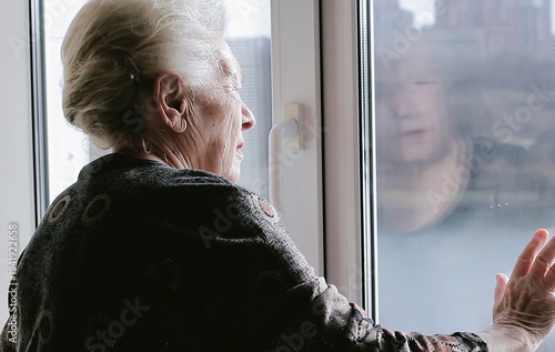 A lonely woman sadly looking out the window, missing her family at the retirement home. Thinking over health problems, loneliness, loss, suffering from apathy, mourning depression.