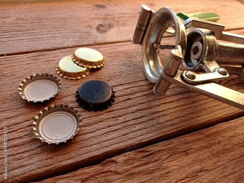 Photography Close up of some crown caps and a manual crown capper on a wooden table