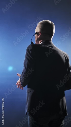 Senior male motivational speaker passionately gestures while delivering an inspiring speech on stage, wearing a headset microphone. Vibrant stage lights illuminate the scene, creating a dynamic and
