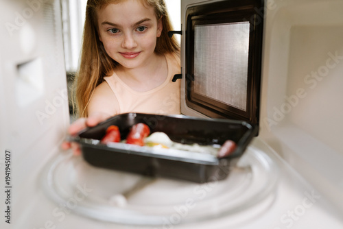 A young girl putting a plastic tray of food into a microwave oven to heat up her meal at home.