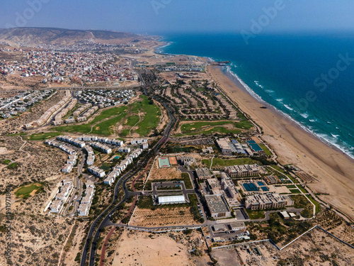 Aerial drone view of the beautiful Taghazout Bay coastline featuring luxury resorts, sandy beaches, lush green golf courses, and the Atlantic Ocean in Agadir, Morocco.