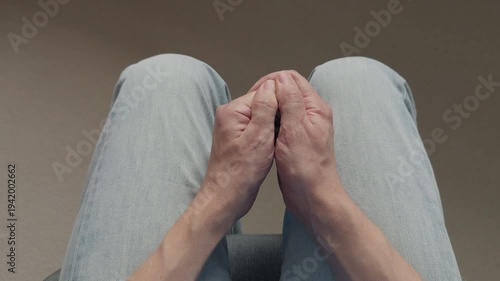 A man sitting in light blue jeans, tightly clenching their hands together in a state of high stress and nervous anxiety. Close up.