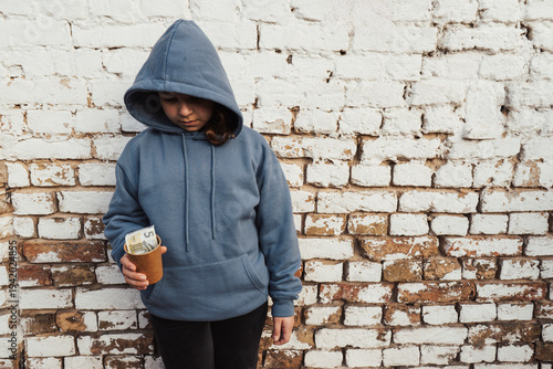 Fotografie A lonely teenage girl in a blue hooded sweatshirt stands by a brick wall, lookin