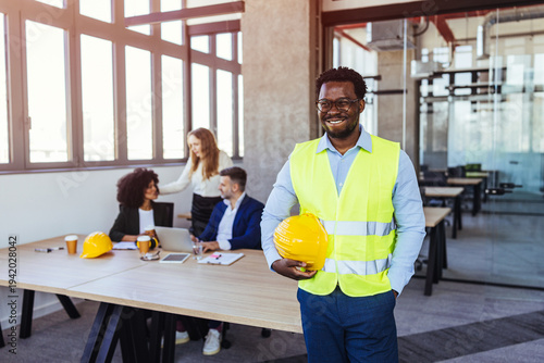 Smiling Construction Engineer in High-Vis Vest Holding Hard Hat in Modern Office Meeting