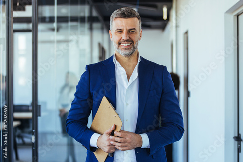 Confident Mature Businessman Smiling in Modern Office Holding Clipboard and Wearing Blue Suit