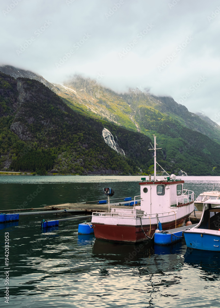 Fototapeta premium Tyssedal fjord atmosphere as small fishing boat anchored by floating dock on calm fjord water with steep mountains and waterfall behind, regional coastal scenery and tourism promotion