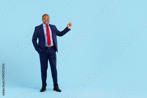 Confident mature black businessman in elegant blue suit pointing aside while standing on clean studio background. Professional african american ceo presenting idea, corporate lifestyle portrait