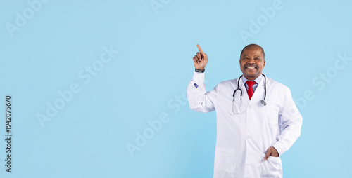 Smiling mature african american doctor pointing upward while standing on blue studio background. Healthcare professional presenting idea, information or medical advice