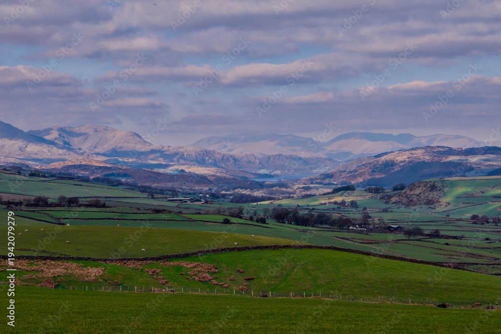 Fototapeta premium Rolling green farmland with stone walls and distant lake and mountains under dramatic cloud cover