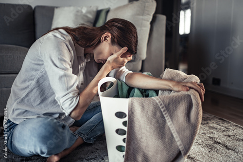 Exhausted woman sitting on floor next to laundry basket feeling overwhelmed by housework, domestic stress and burnout concept
