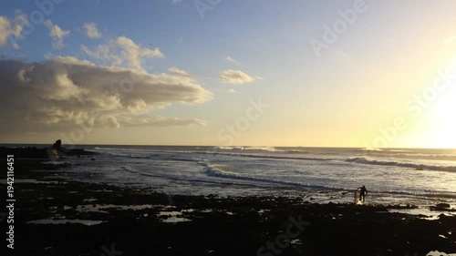 Surfer walking out of the ocean with a small shortboard during golden hour on Tenerife, illuminated by warm sunset light along the Atlantic coastline of the Canary Islands
