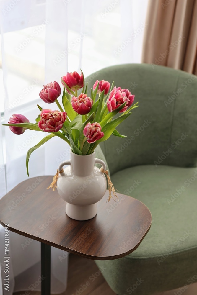 Obraz premium A beautiful bouquet of pink double tulips in a white ceramic vase on a wooden side table by a green armchair. Concept of cozy spring home decor.