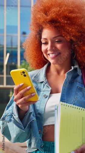 Latin student girl using cellphone phone standing at university campus. Smiling young african american woman with curly ginger hair using smartphone mobile app for studying outdoors. Vertical