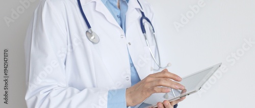 Photos Unknown female doctor, wearing white coat over a blue blouse and a stethoscope, is using a digital tablet, suggesting innovative healthcare practices in medicine